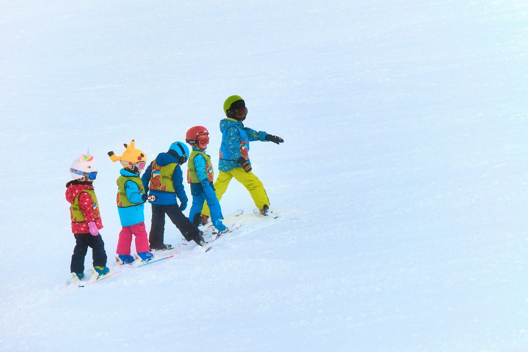 Five small children in bright helmets and coloured bibs standing in a line on skis in the snow